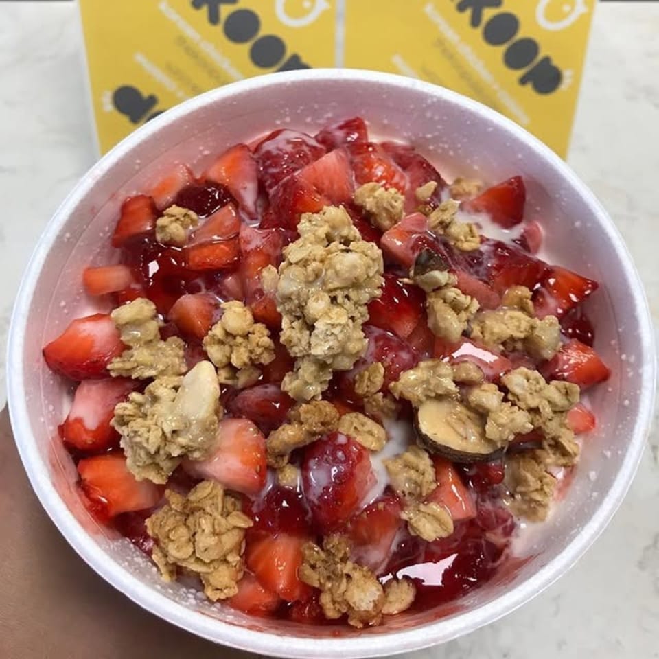 A white bowl filled with sliced strawberries, red sauce, granola, and a drizzle of white cream.