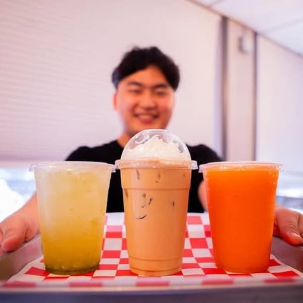 Smiling person holding a tray with three drinks: yellow, iced coffee with whipped cream, and orange.