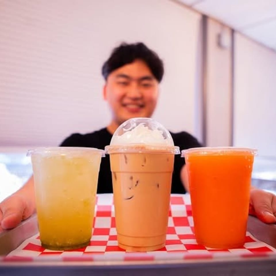 Smiling person holding a tray with three drinks: yellow, iced coffee with whipped cream, and orange.