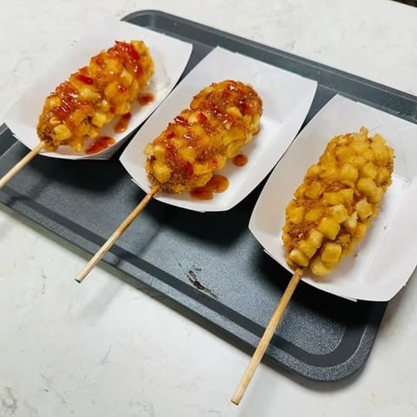 Three Korean corn dogs, two drizzled with sauce, on a black tray.