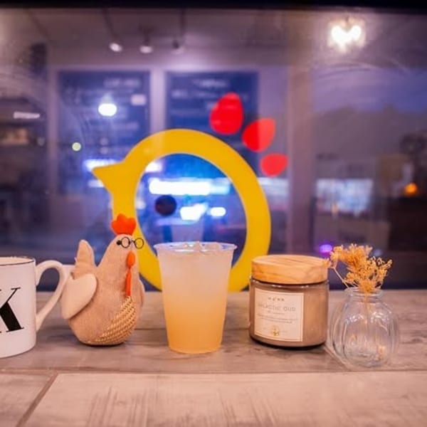 A chicken figurine, drink, and candle on a wooden table with The Koop logo in the background.