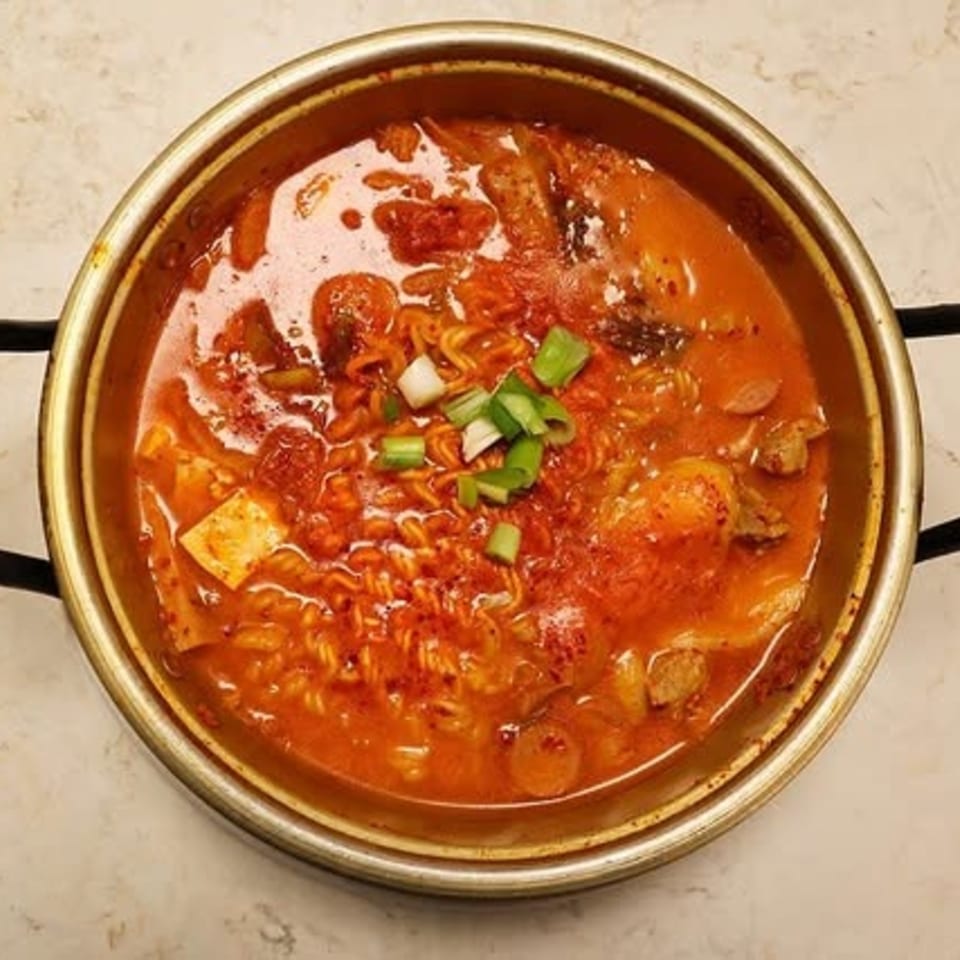 A golden pot of Korean budae jjigae, or army stew, with ramen, tofu, and green onions.