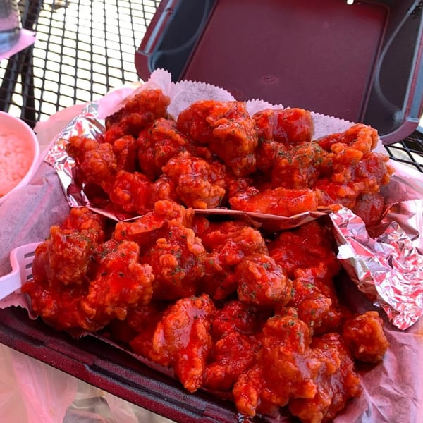A black takeout container filled with bright red, saucy Korean fried chicken pieces, garnished with herbs.
